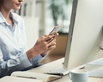 Unrecognizable Woman Sitting In Office In Front Of Computer And Using Smartphone